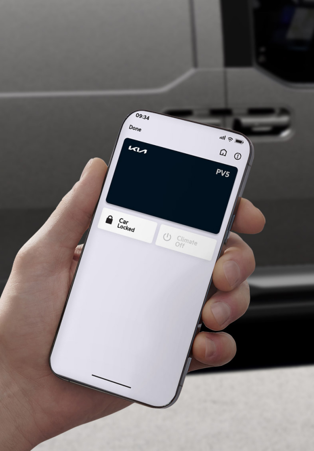 Hand hält Smartphone mit angezeigtem digitalen Schlüssel des Kia PV5 vor dem Fahrzeug. 